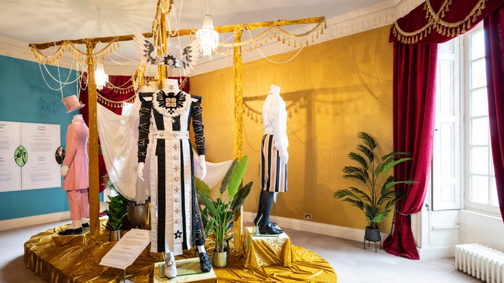 A view of the exhibition room for All Dressed Up at Plas Newydd, with an opulent display of 5 costumes from the film Madfabulous in the centre and rich red velvet curtains and gold walls.